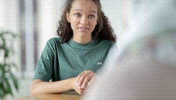 Eine Jugendliche ist im Gespräch mit einer anderen Person am Tisch | © FatCamera - Getty Images/iStockphoto